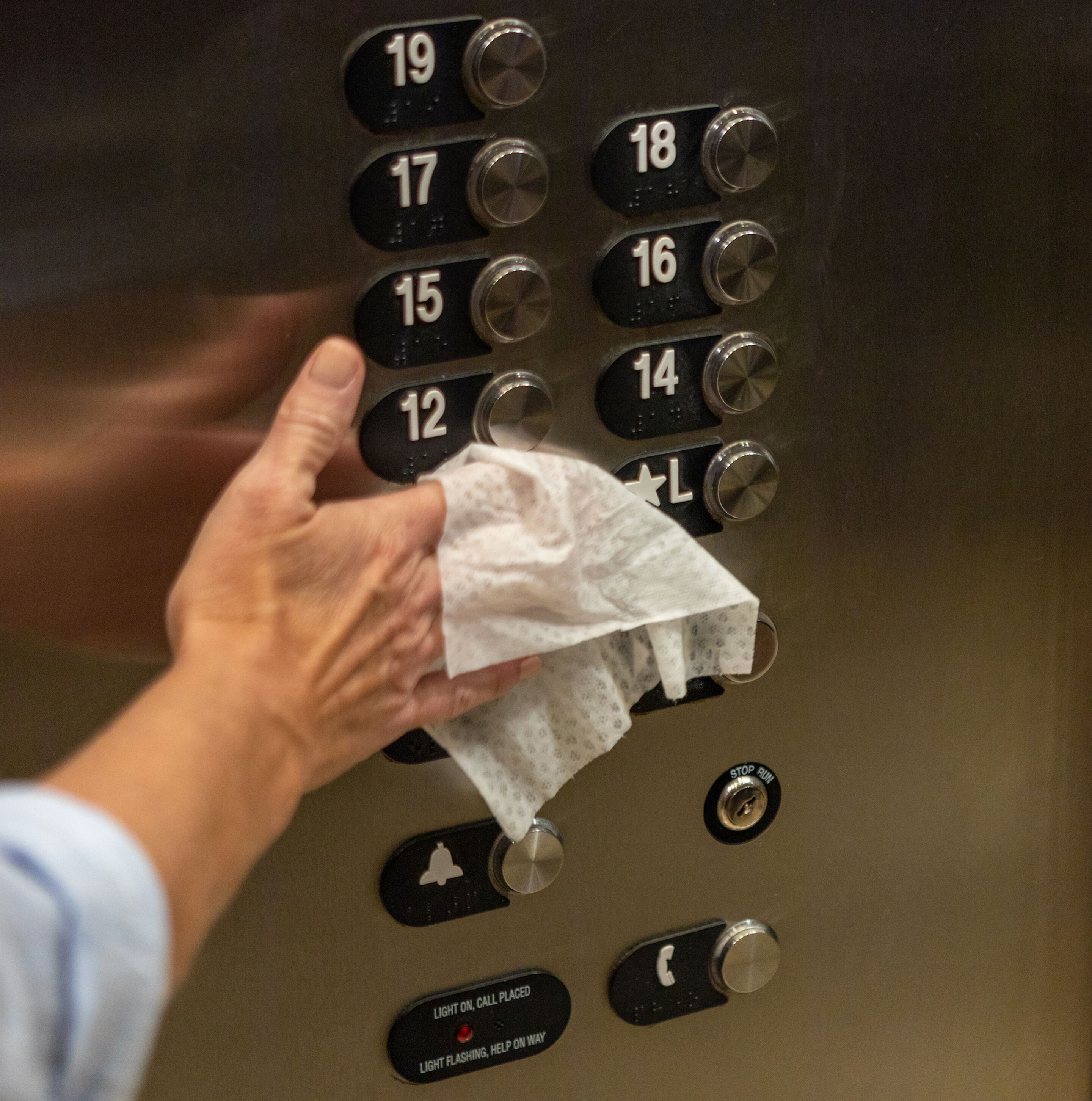 Een hand houdt een zakdoek vast nabij een bedieningspaneel van een lift, met genummerde en noodknoppen.