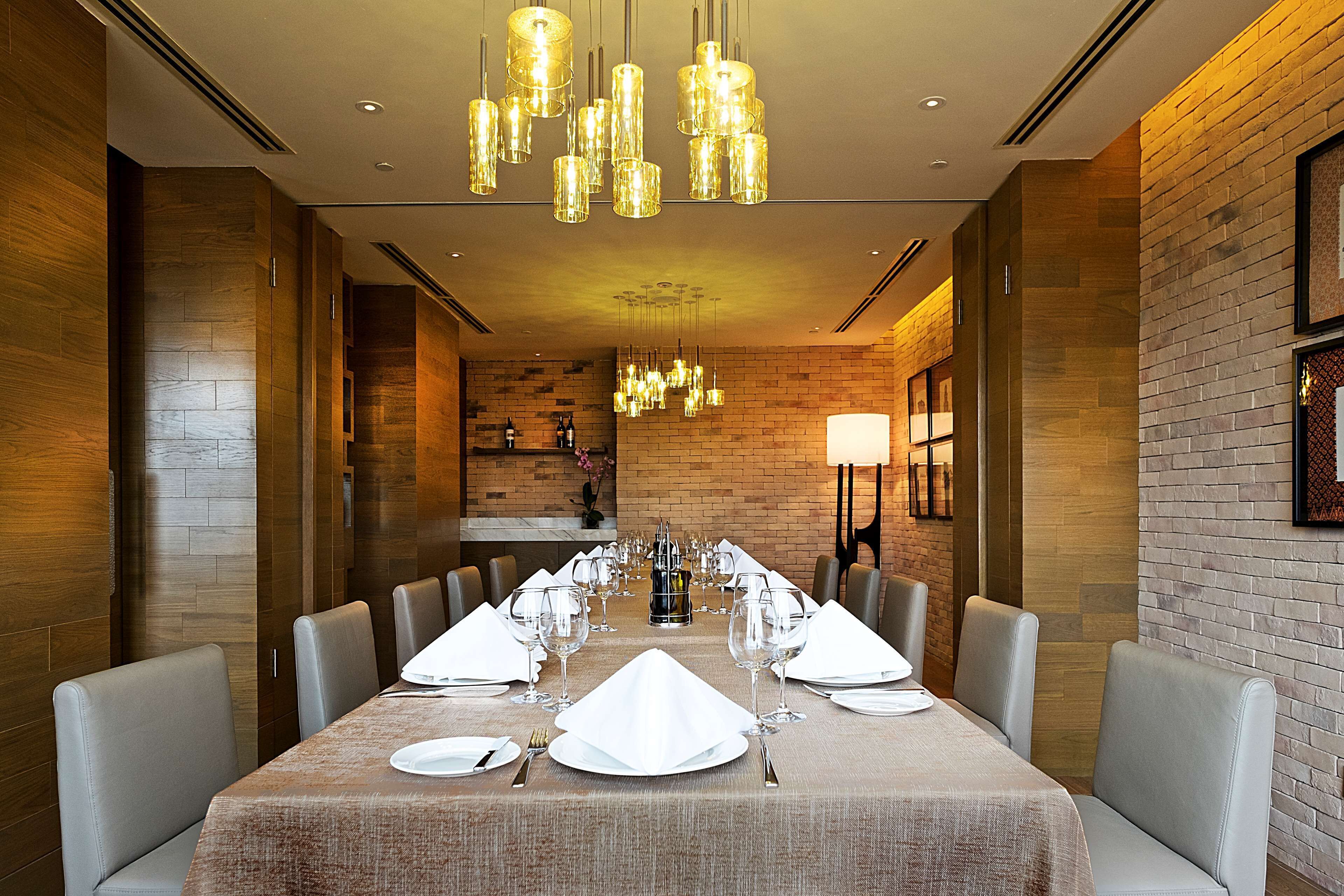 A dining area with a long table set for a meal, surrounded by cushioned chairs, and illuminated by multiple hanging light fixtures.