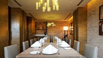 Uma sala de jantar com uma mesa comprida posta para refeição, rodeada por cadeiras estofadas, e iluminada por várias luminárias pendentes.
