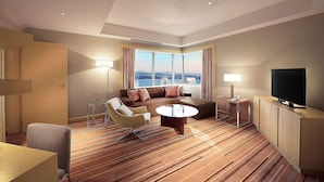 Habitación de hotel moderna con un amplio sofá, una mesa de centro, televisor y vistas a la ciudad a través de grandes ventanales.