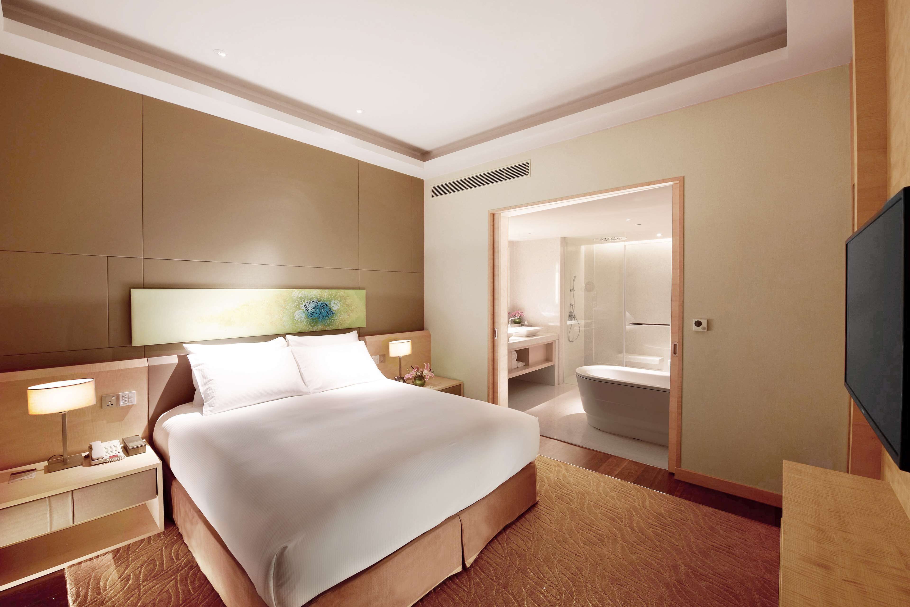 A modern hotel room with a large bed, bedside tables, a flat-screen TV, and a bathroom visible through an open door.