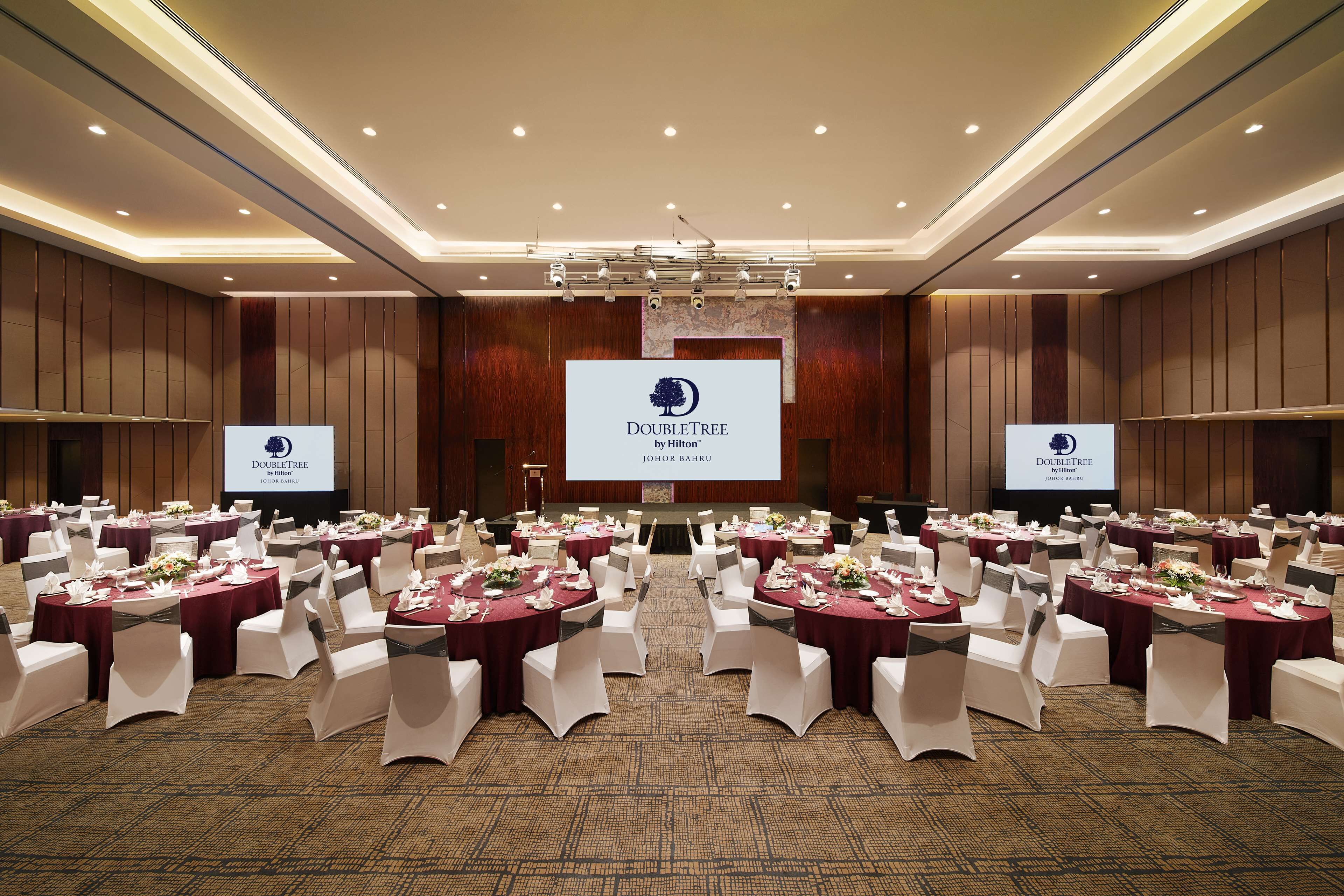 Un salón de conferencias con mesas redondas, sillas blancas y una pantalla grande que muestra el logotipo de DoubleTree by Hilton.