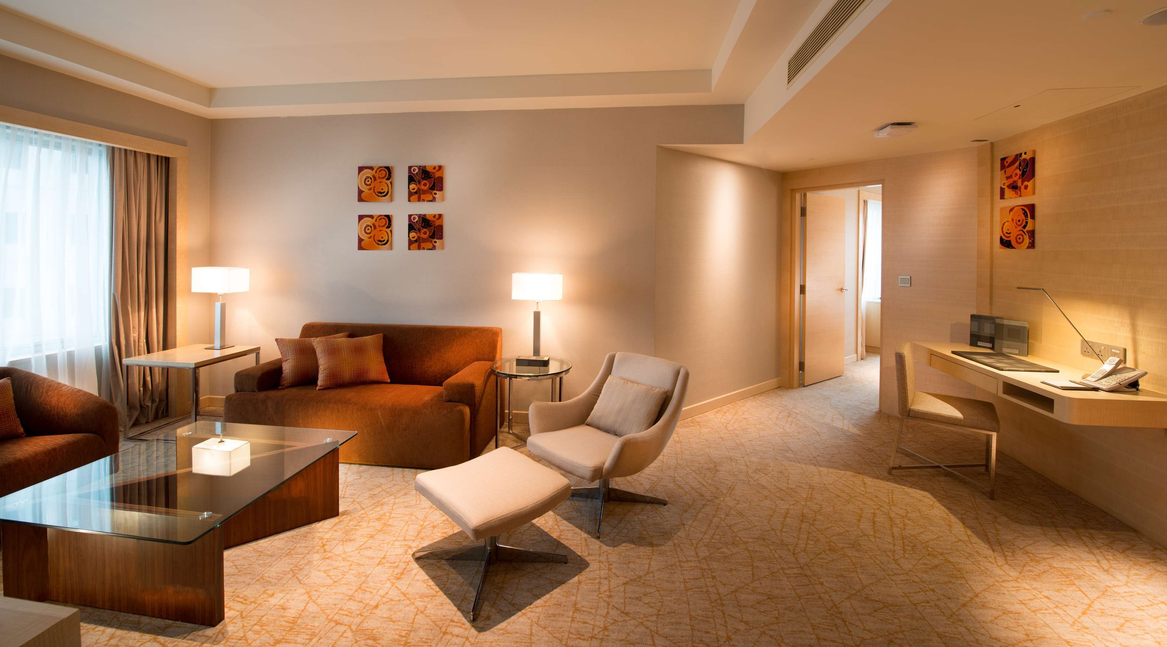 Una camera d'albergo moderna con un divano, una poltrona e un tavolino in vetro. C'è una scrivania con una lampada e un telefono. Il pavimento è rivestito di moquette e le pareti sono decorate con opere d'arte.