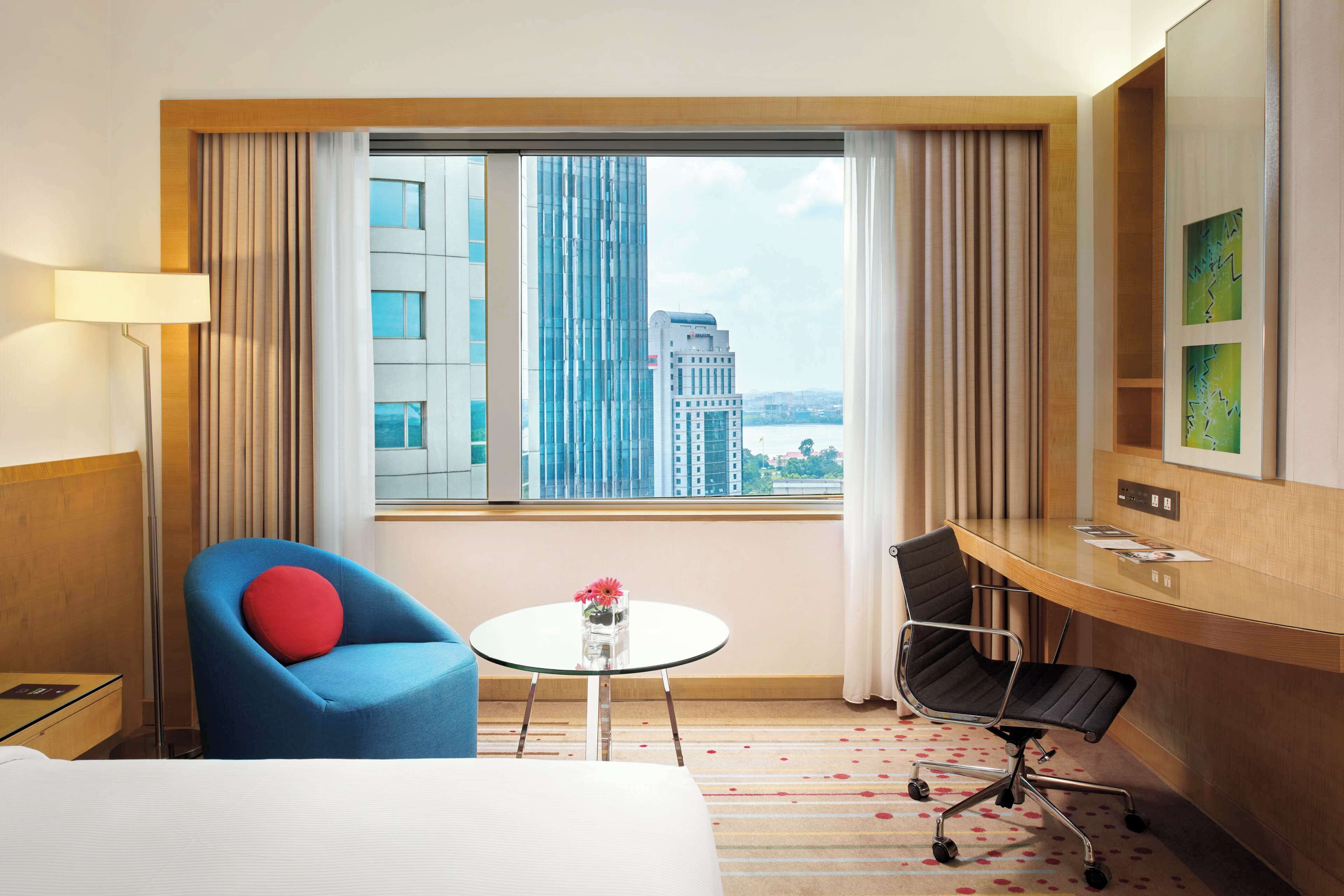 Ein Hotelzimmer mit einem Bett, einem blauen Sessel mit rotem Kissen, einem Schreibtisch mit Stuhl, einem kleinen Tisch mit Blumenvase und einem großen Fenster mit Blick auf die Stadt.