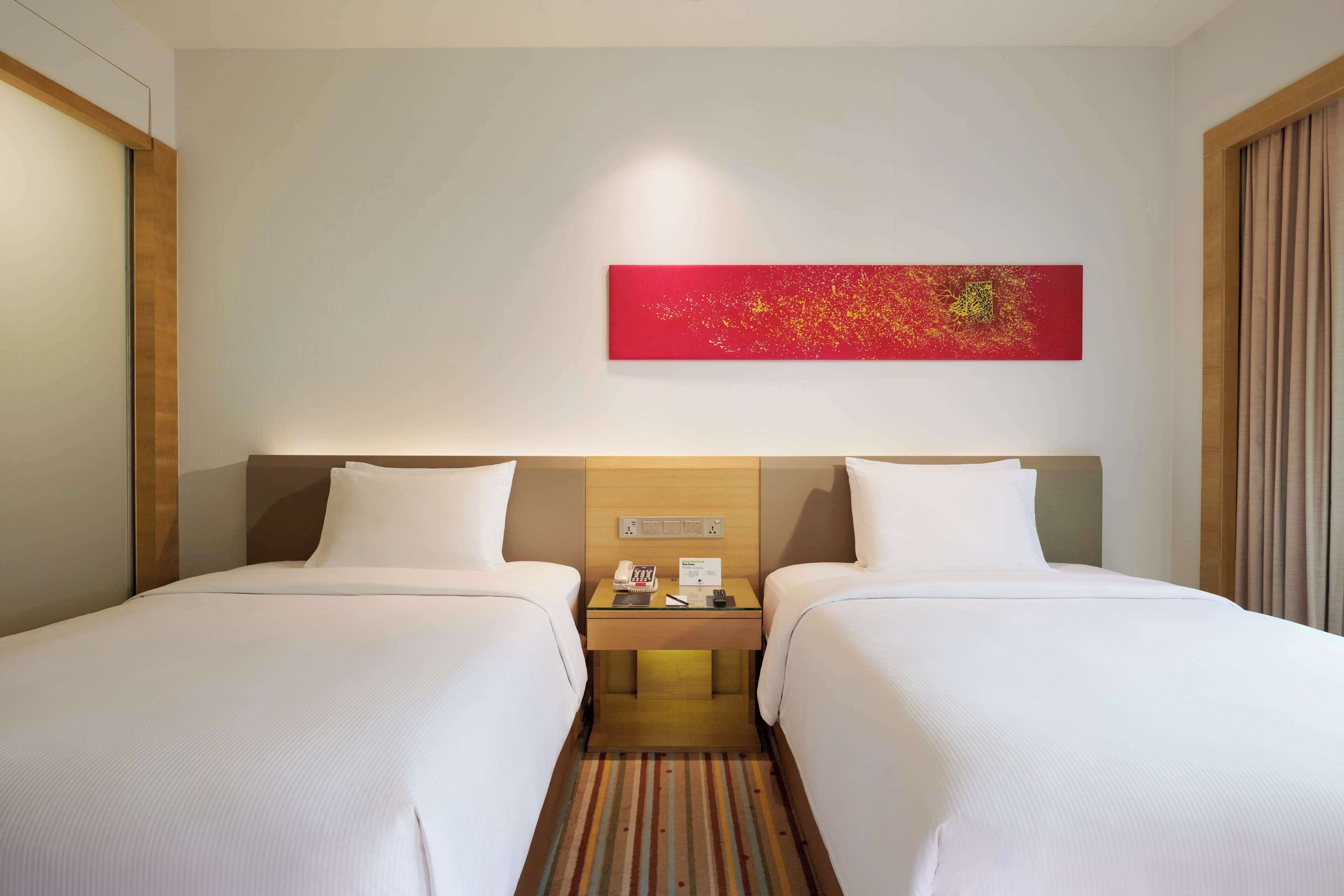A hotel room with two beds, a nightstand with a phone and a remote control, a red abstract painting on the wall, and a striped carpet.