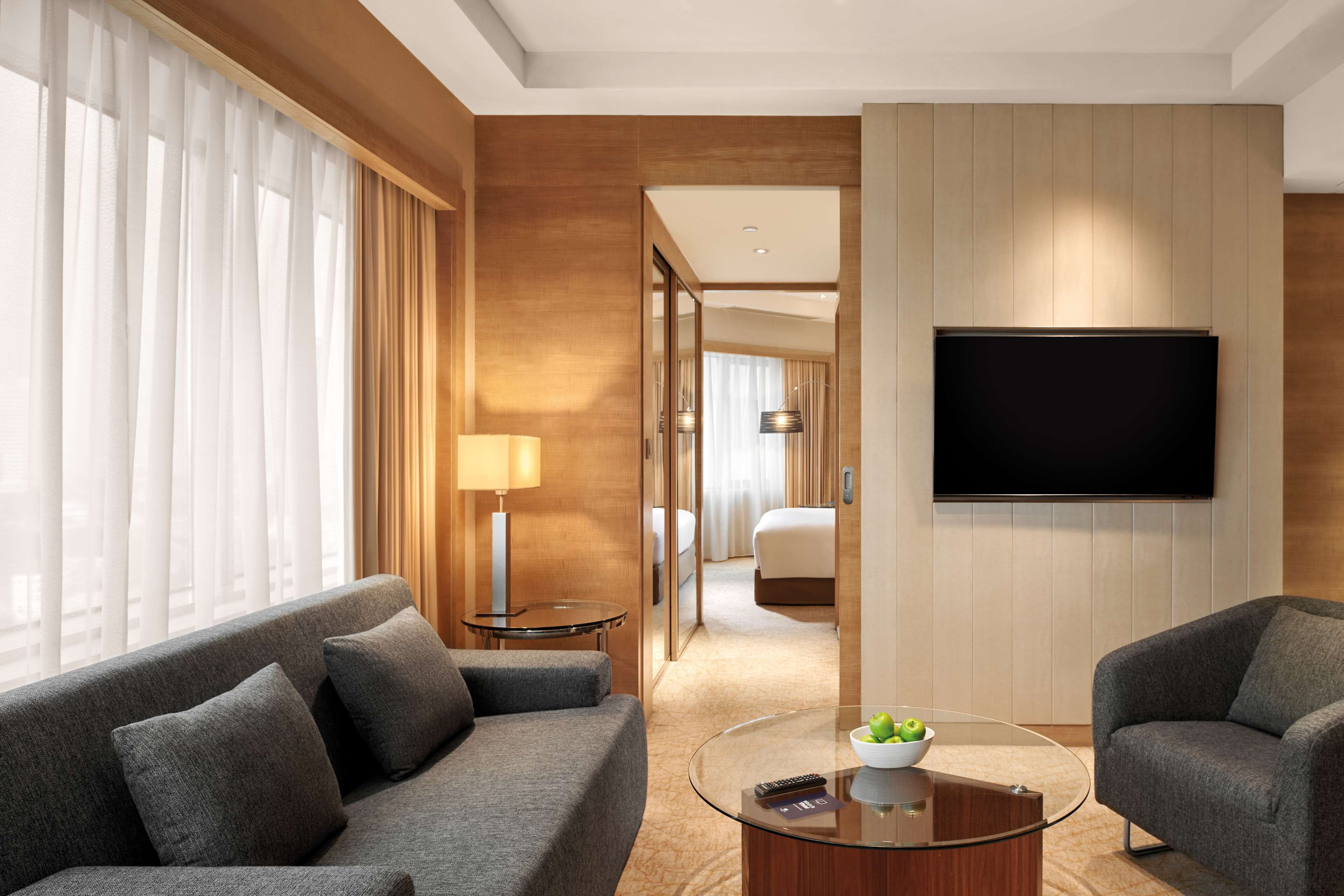Quarto de hotel moderno com um sofá cinzento, uma mesa de centro de vidro, uma televisão de ecrã plano e vista para o corredor.