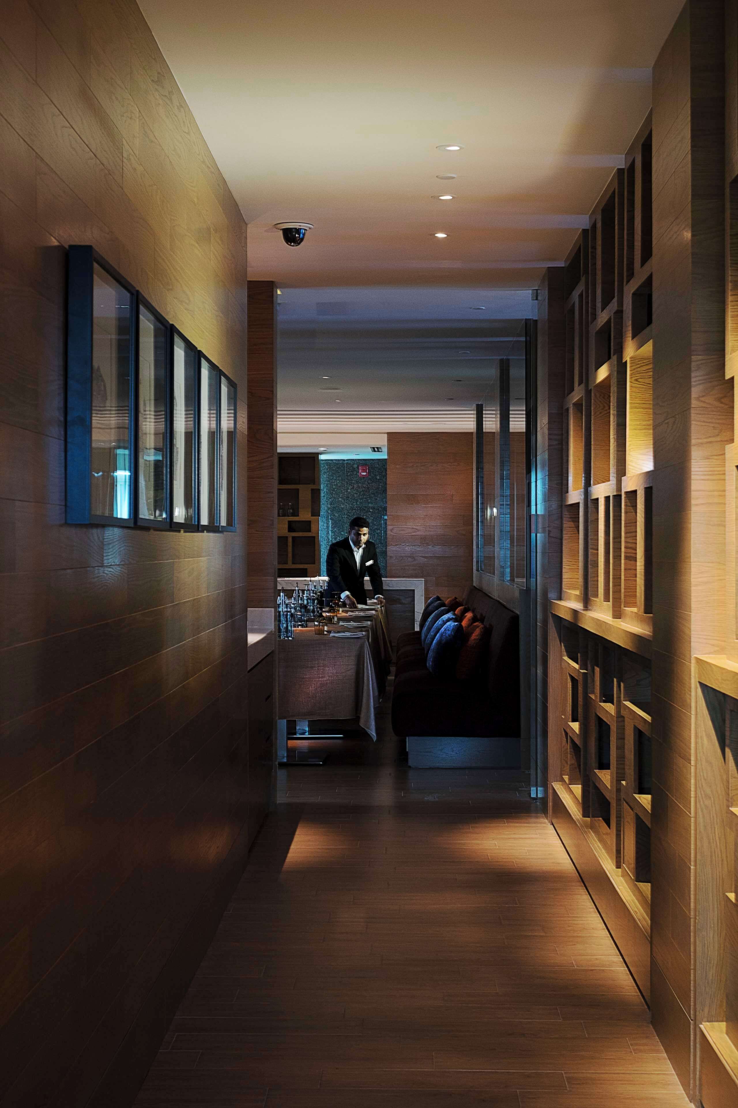 A modern hotel corridor with wooden paneling, a reception desk, a seating area, and a security camera on the ceiling.
