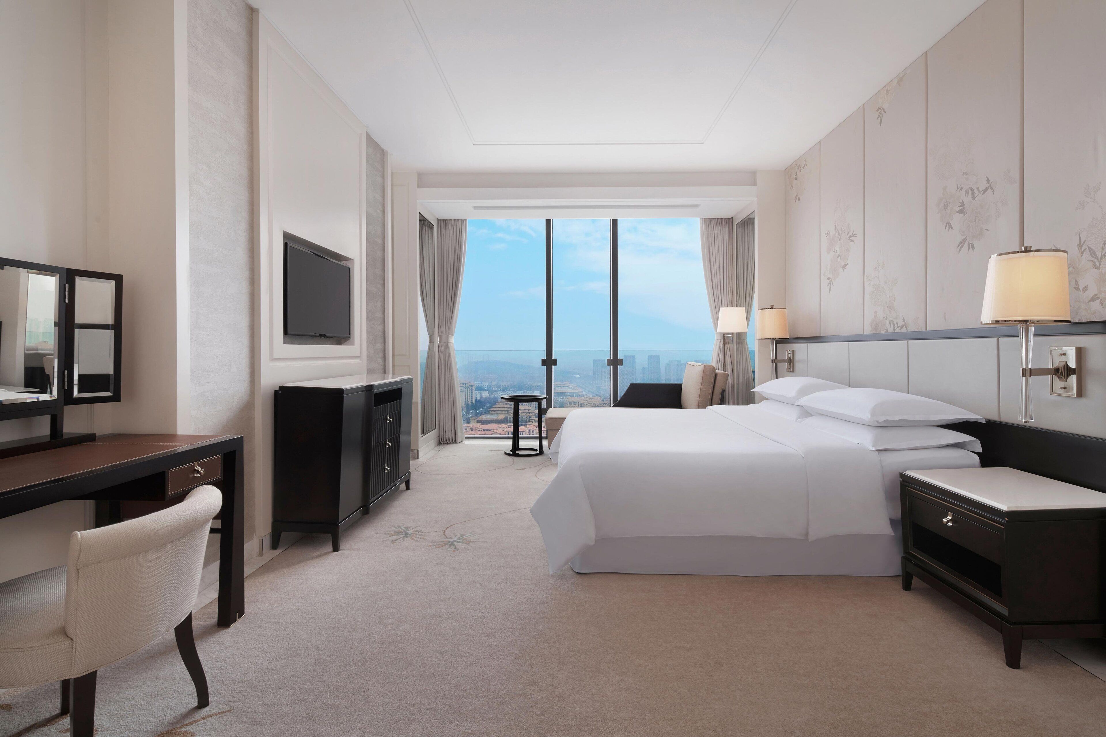A hotel room with a large bed, a desk with a chair, a TV, and a view of the city through large windows.