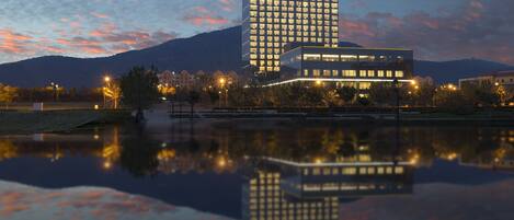A modern hotel with a reflection in a body of water, surrounded by trees and a mountain in the background.