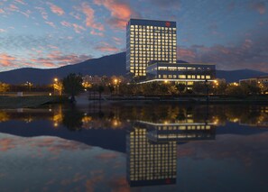 A modern hotel with a reflection in a body of water, surrounded by trees and a mountain in the background. - Sheraton Qingdao Licang Hotel (Qingdao)