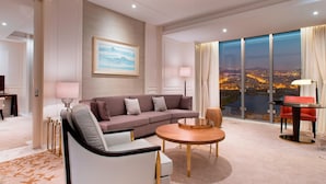 Une chambre d’hôtel moderne dotée d’une grande fenêtre donnant sur la ville, d’un canapé, d’une table basse ronde et d’un coin repas avec une chaise rouge.