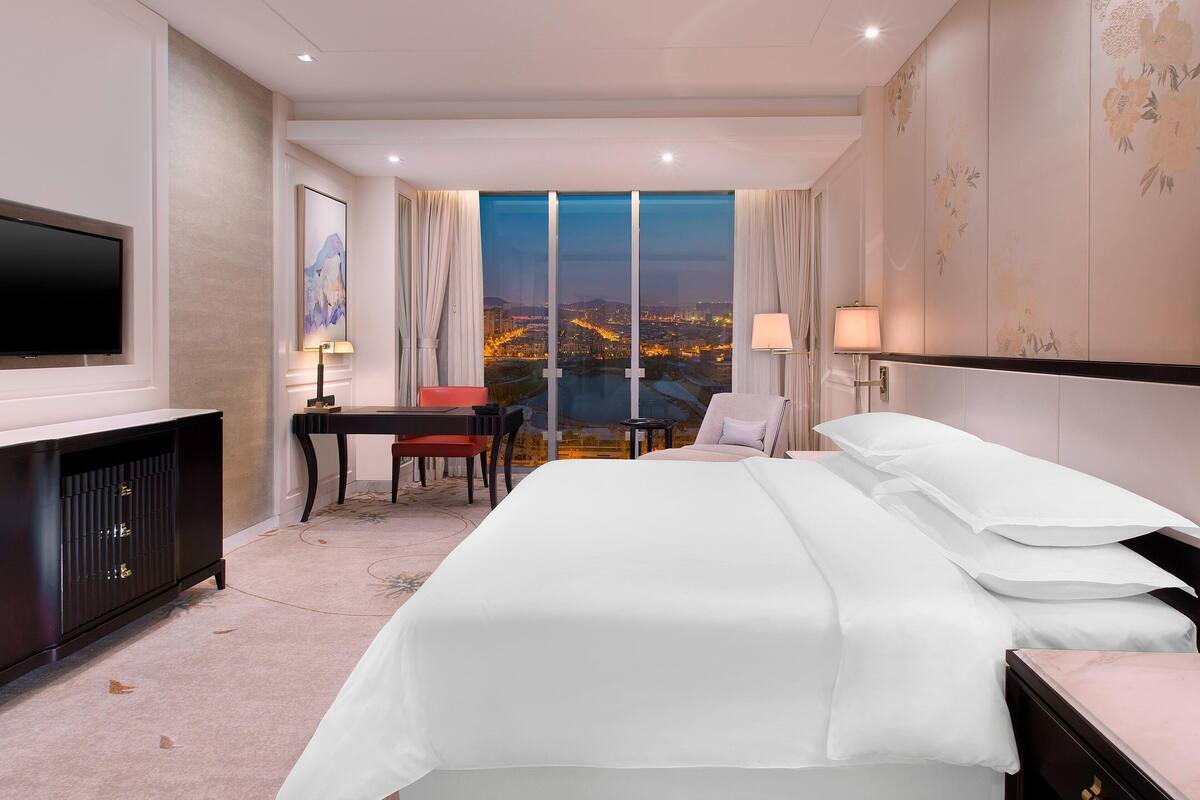 Quarto de hotel com uma cama grande, uma escrivaninha com cadeira, uma televisão e vista para a paisagem urbana ao entardecer.