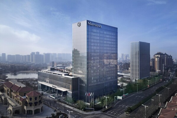 A modern Sheraton hotel with a glass facade, situated in a cityscape with other high-rise buildings and a river in the background. - Sheraton Qingdao Licang Hotel (Qingdao)