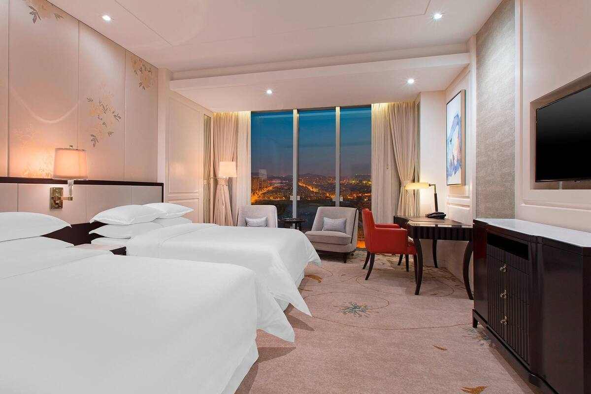 Quarto de hotel com duas camas, uma escrivaninha, uma cadeira, televisão e uma janela grande com vista para a cidade.
