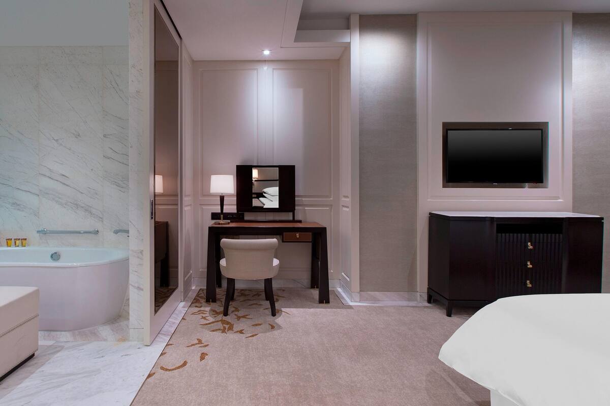 Quarto de hotel moderno com banheiro de mármore, escrivaninha com cadeira, televisão e guarda-roupa.