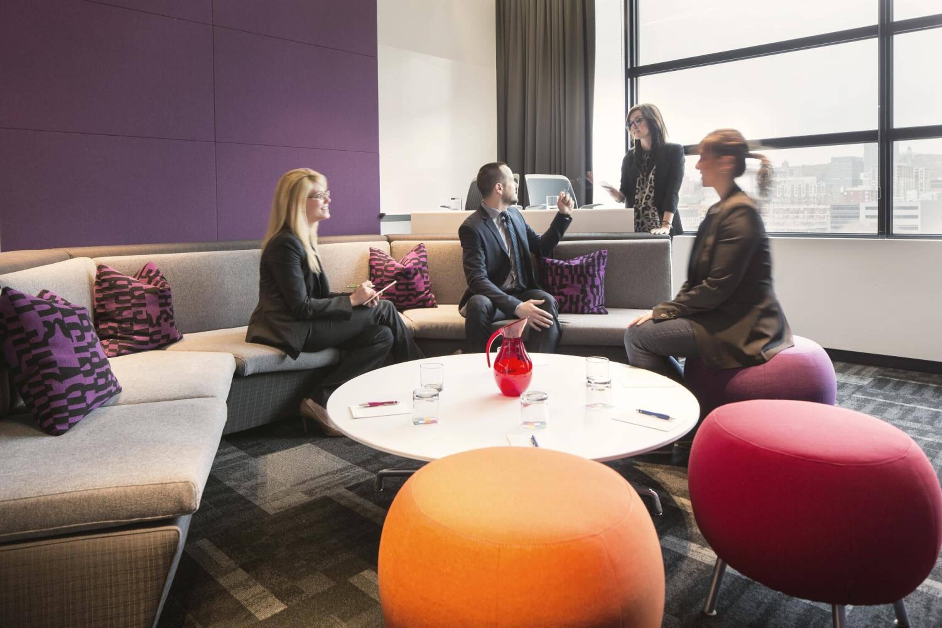 A modern meeting room with a sofa, ottoman, and coffee table, featuring a city view through large windows.