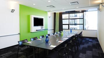 A conference room with a long table, chairs, a flat-screen TV, and a large window with a view of buildings.