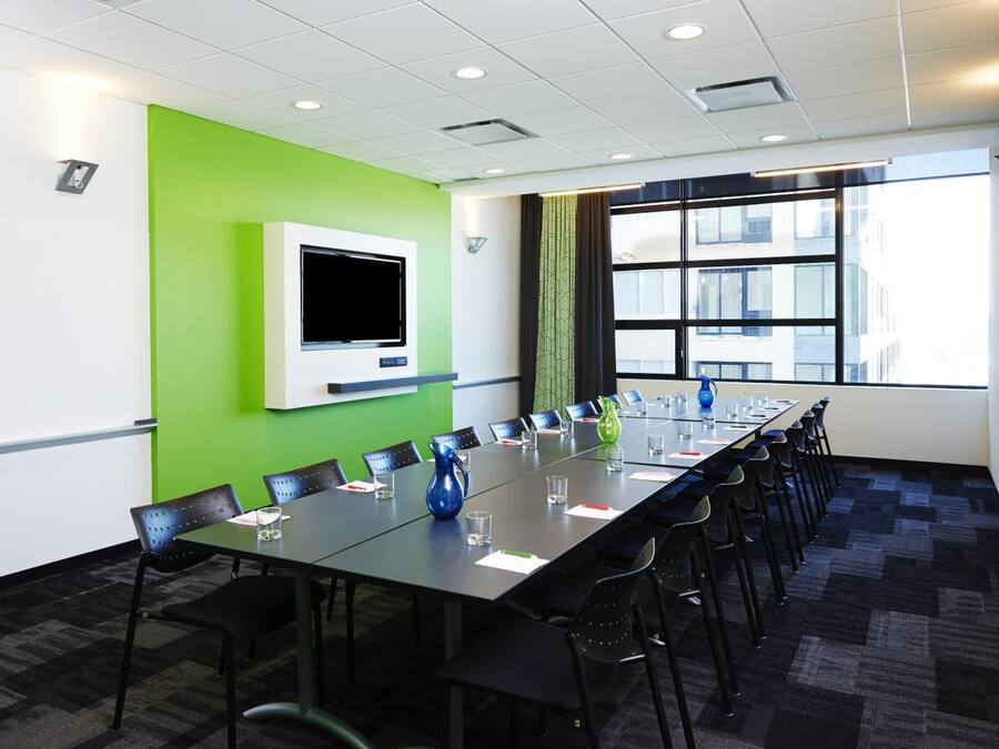 A conference room with a long table, chairs, a flat-screen TV, and a large window with a view of buildings.