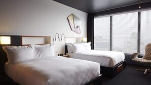 A hotel room with two beds, a modern chair, a wall-mounted lamp, and a window with blinds. - Hôtel Alt Montréal (Montreal)