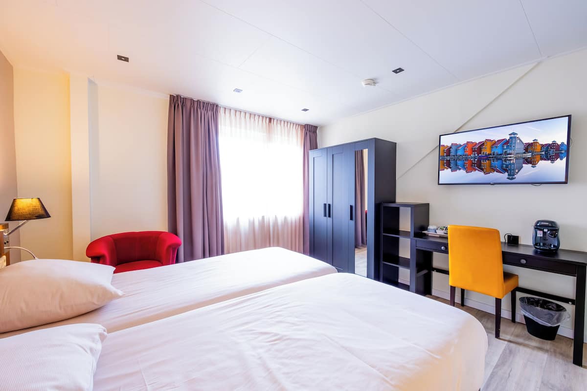 A hotel room with two beds, a desk, a chair, a wardrobe, and a painting on the wall.