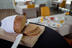 A loaf of bread on a wooden cutting board with a knife, a dining table with plates and a glass of orange juice, and a modern interior with yellow chairs and a white tablecloth. - Best Western Plus Hotel Restaurant Aduard (Aduard)