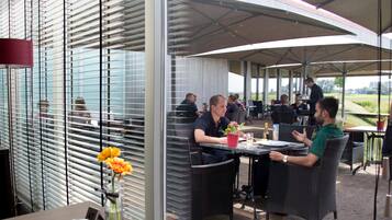 An outdoor dining area with wicker chairs and tables, a covered section with umbrellas, and a view of the landscape through large windows.