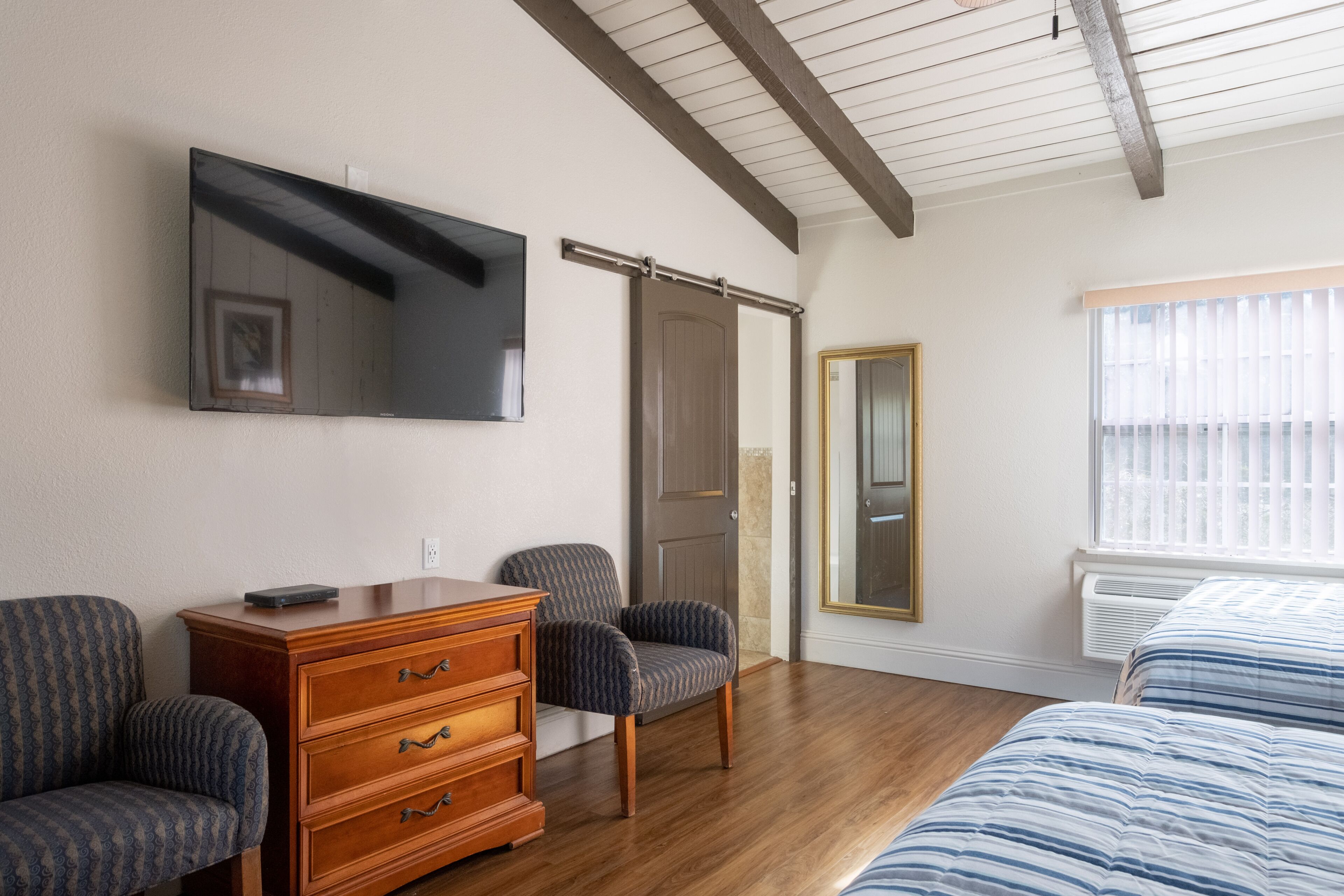 A bedroom with a wooden dresser, two patterned chairs, a bed with striped bedding, a flat-screen TV mounted on the wall, and a sliding door.