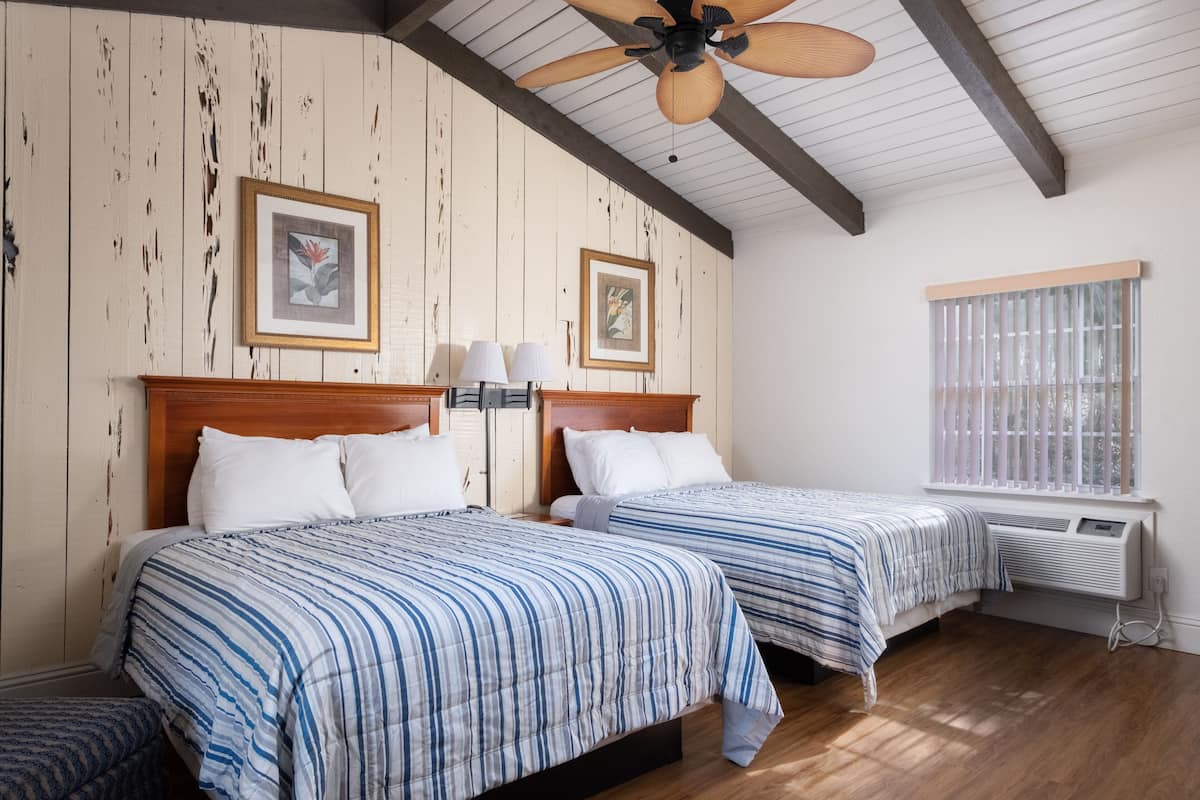 A bedroom with two beds, wooden beams on the ceiling, framed artwork on the walls, and a window with blinds.