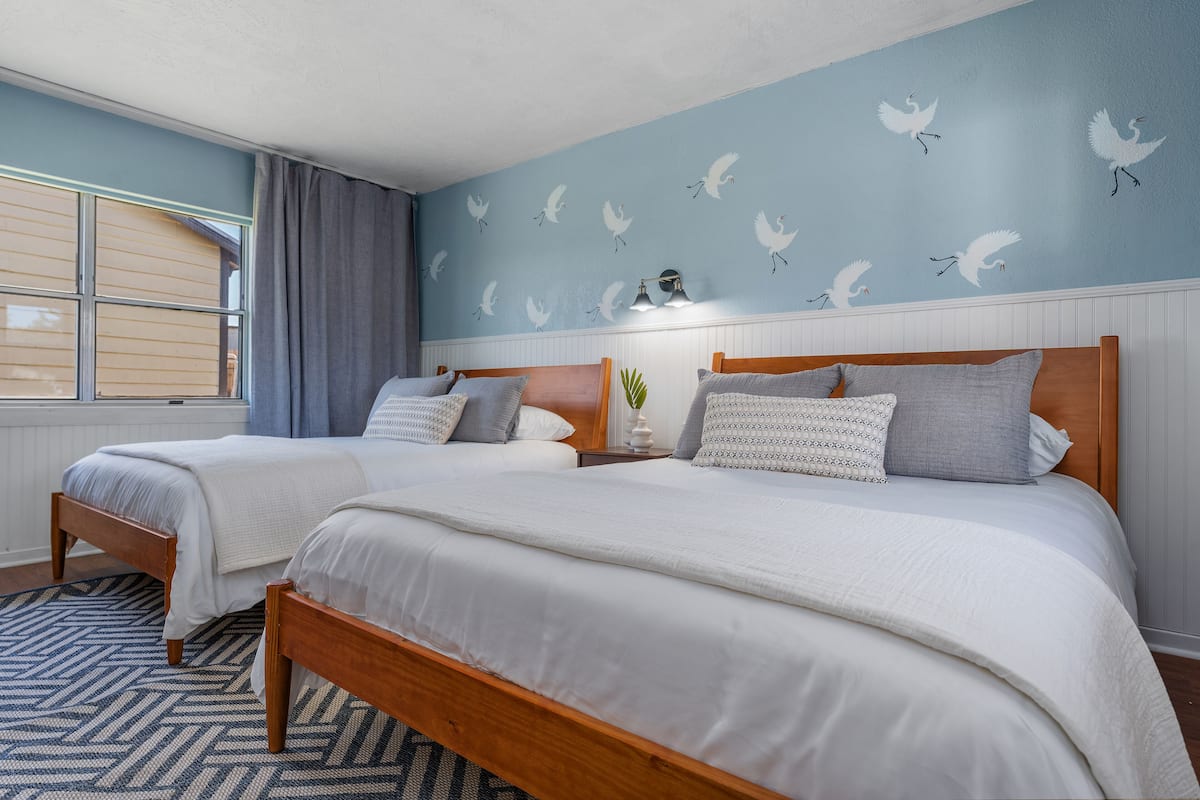A bedroom with two beds, a patterned rug, a window with curtains, and a wall with bird decals.