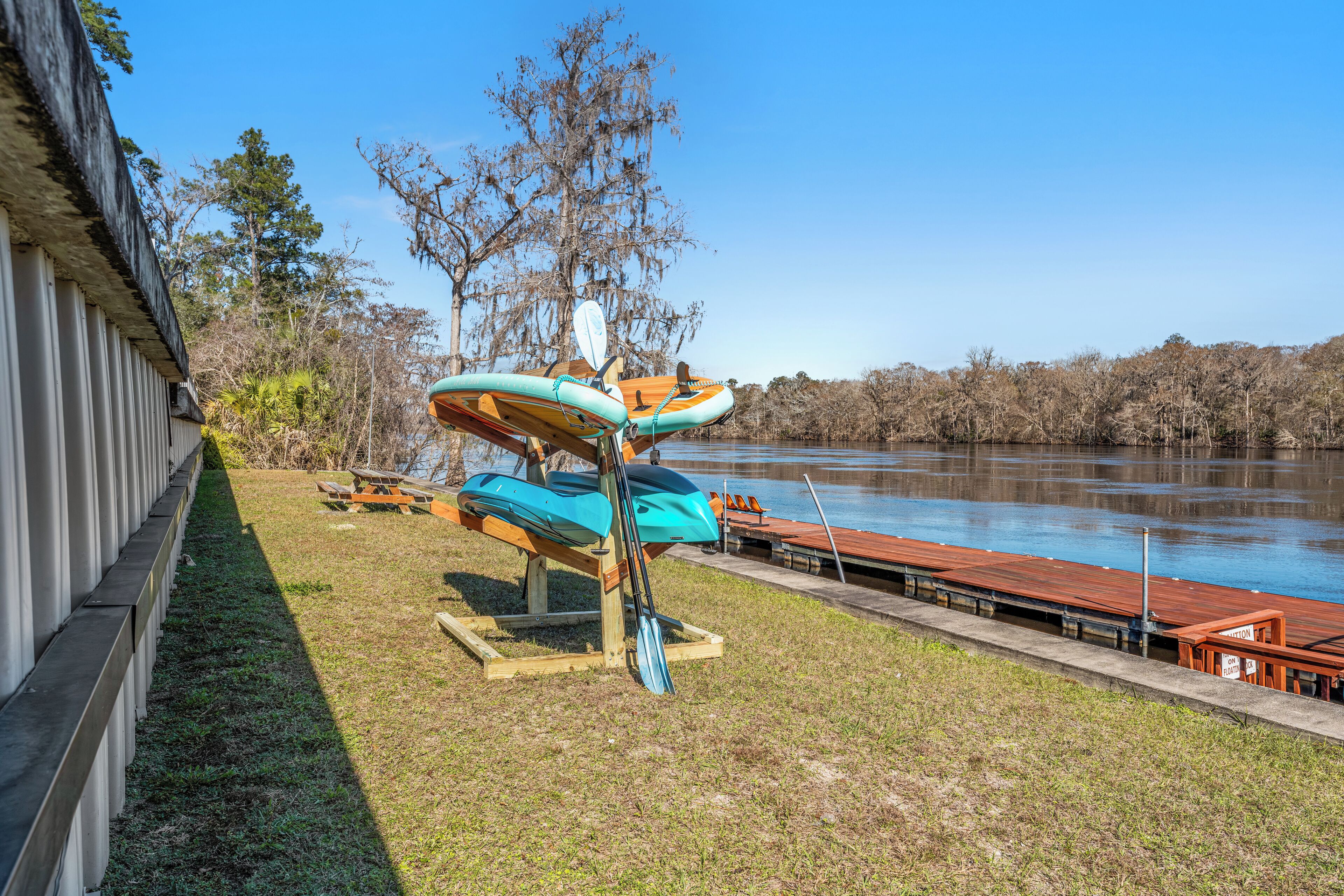 Un paisaje junto al lago con kayaks y una tabla de remo, un muelle de madera y una zona cubierta de césped.