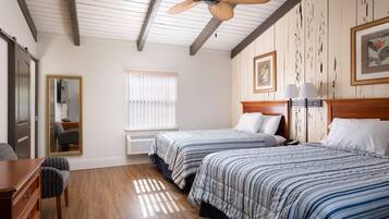 Habitación de hotel con dos camas, pisos de madera, ventilador de techo, ventana con persianas, espejo, escritorio y una silla.
