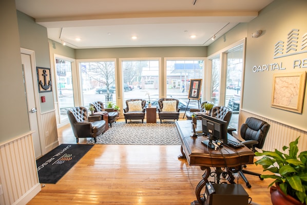 A well-lit room with a wooden floor, leather sofas, a desk with a computer, and large windows offering a view of the street. - 84 Main  (Kennebunk)