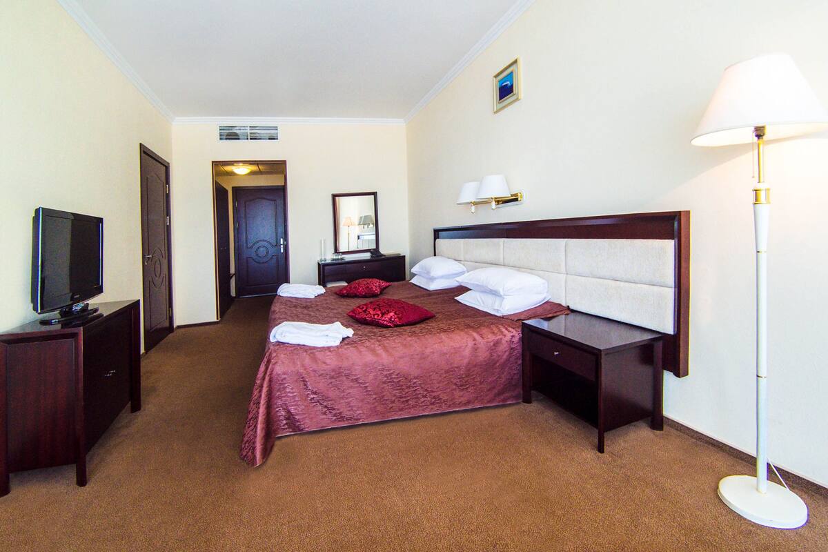 Ein Hotelzimmer mit einem großen Bett, einem Fernseher auf einem Standfuß, einem Nachttisch, einer Lampe und einer Tür zu einem anderen Raum.