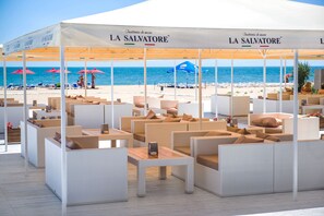 A beachside outdoor seating area with white wicker furniture and red umbrellas, under a canopy with "LA SALVATORE" branding. - Black Sea Bugaz (Zatoka)