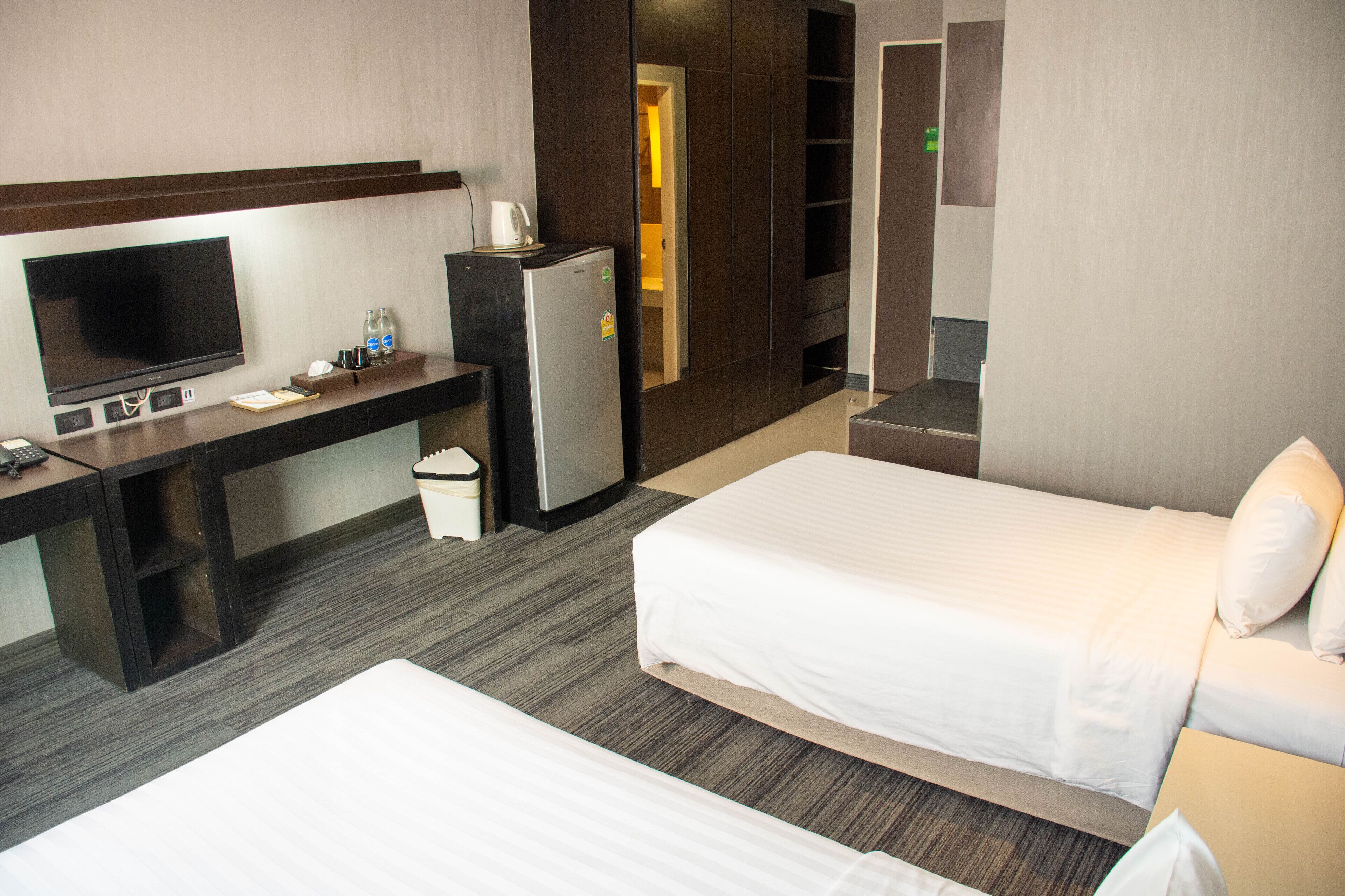 A hotel room with a bed, a desk with a TV, a refrigerator, and a trash bin.