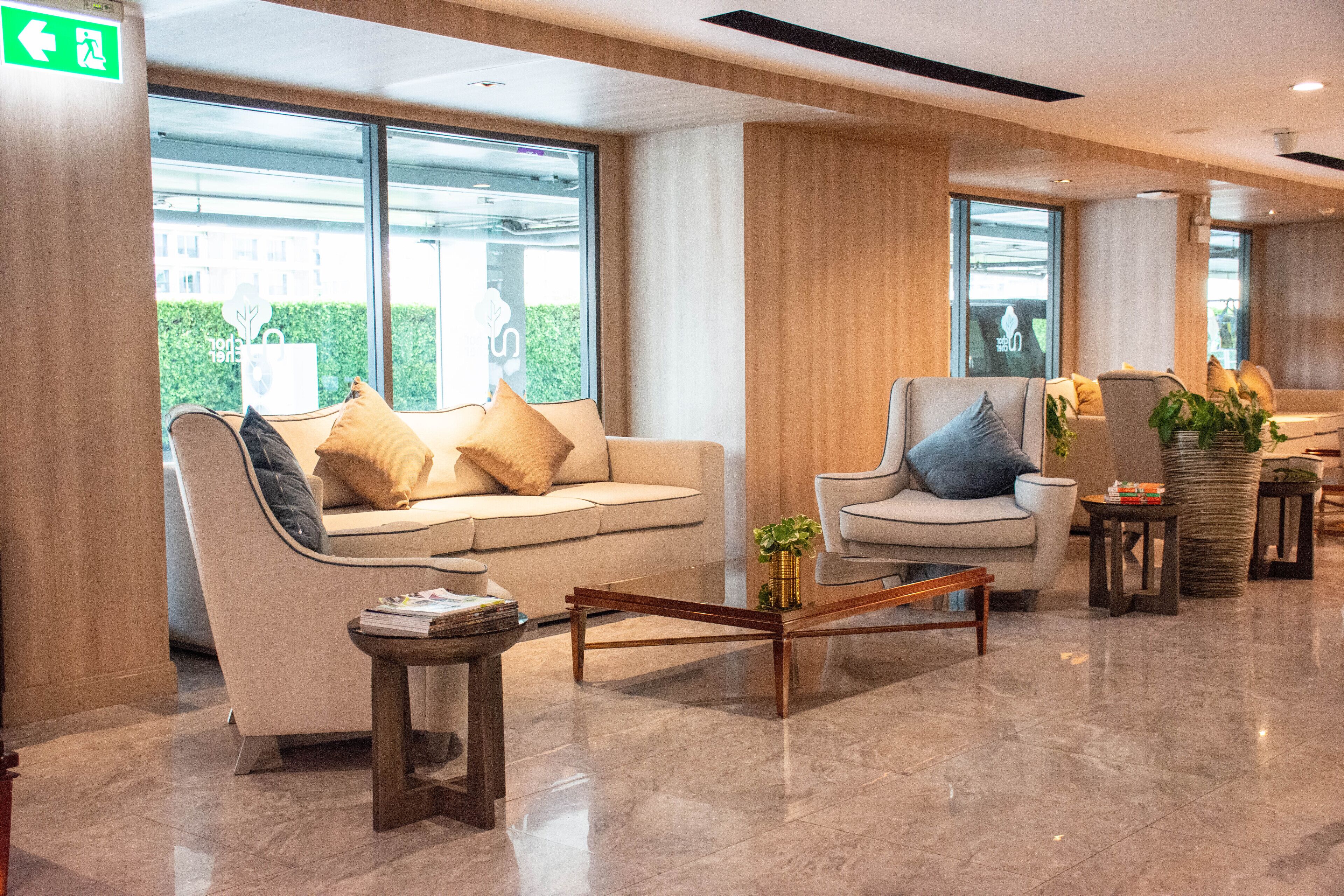 A modern lobby with a sofa, armchair, and coffee table, large windows with a view, and a green exit sign.