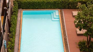 An aerial view of a rectangular swimming pool surrounded by a brick deck, with a hedge and a tree on one side.