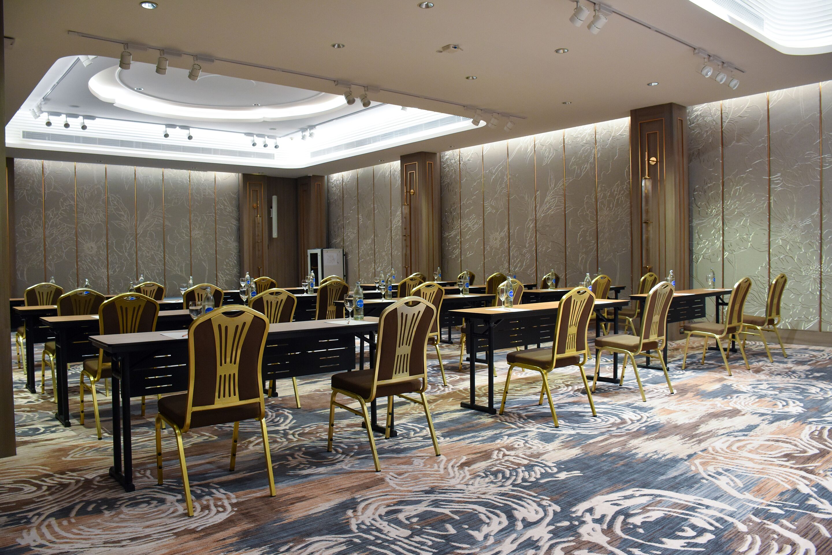 A conference room with rows of tables and chairs, a large ceiling light, and patterned walls.