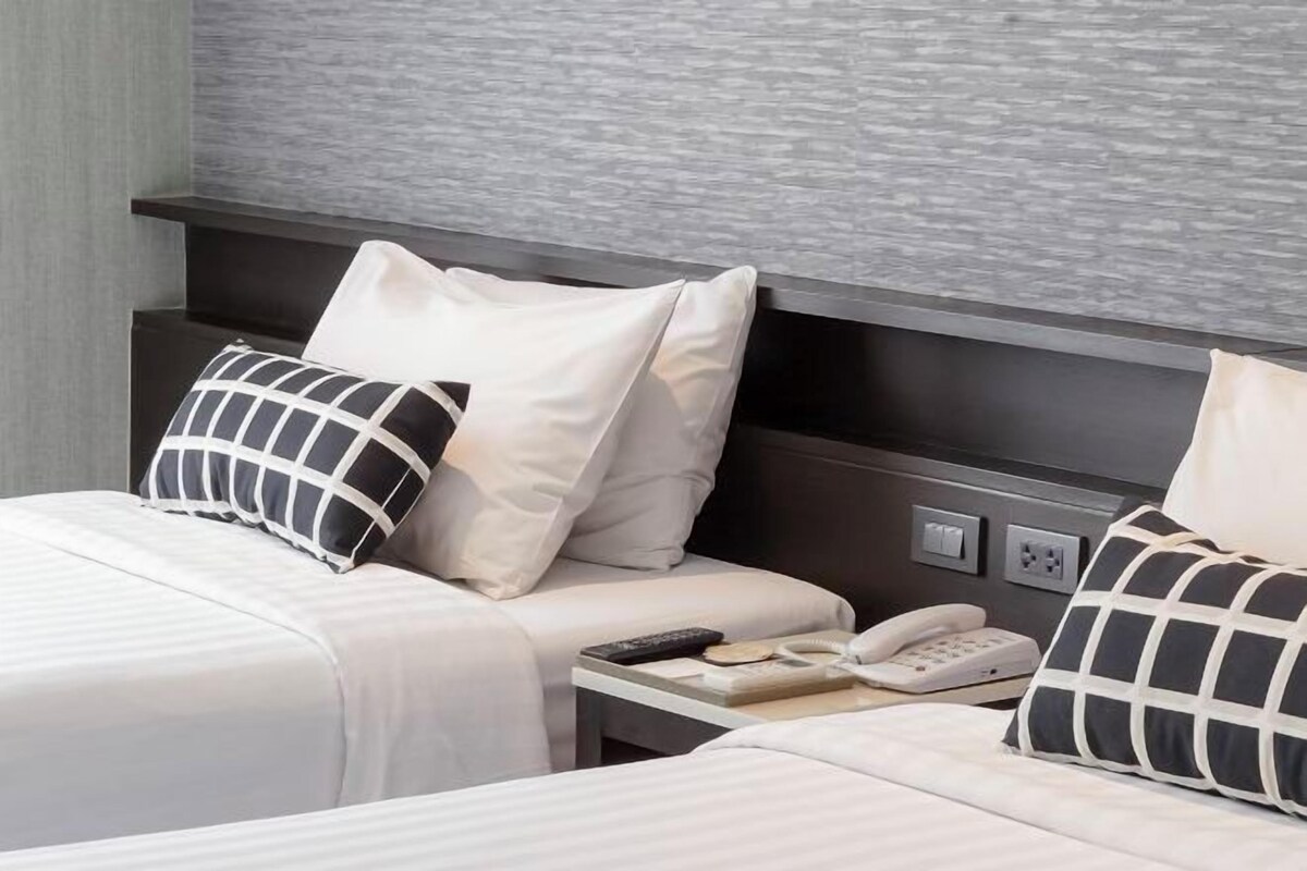 A hotel room with two beds, each with white bedding and black and white patterned pillows. There is a bedside table with a phone and a remote control.