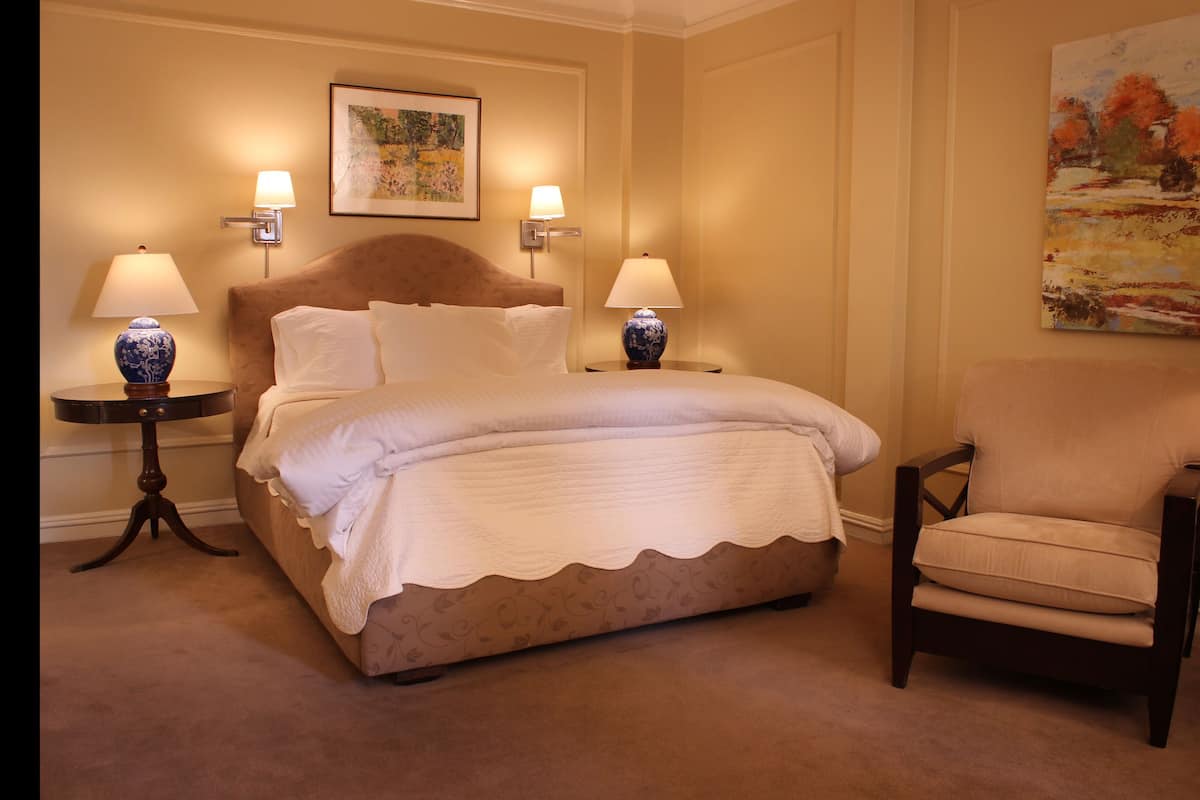 A neatly arranged bedroom with a large bed, two bedside tables with lamps, a chair, and two paintings on the walls.