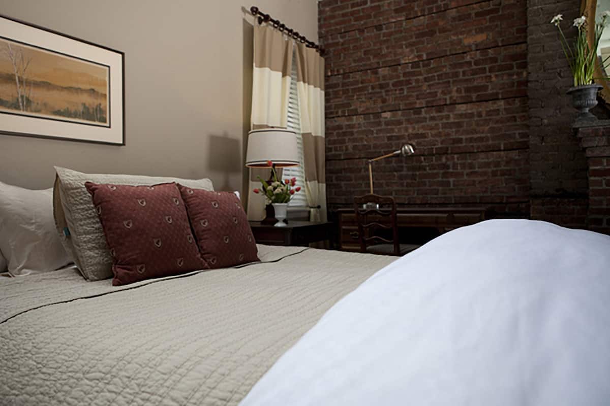 A neatly made bed with white and red pillows, a framed landscape painting on the wall, a lamp, and a brick wall.