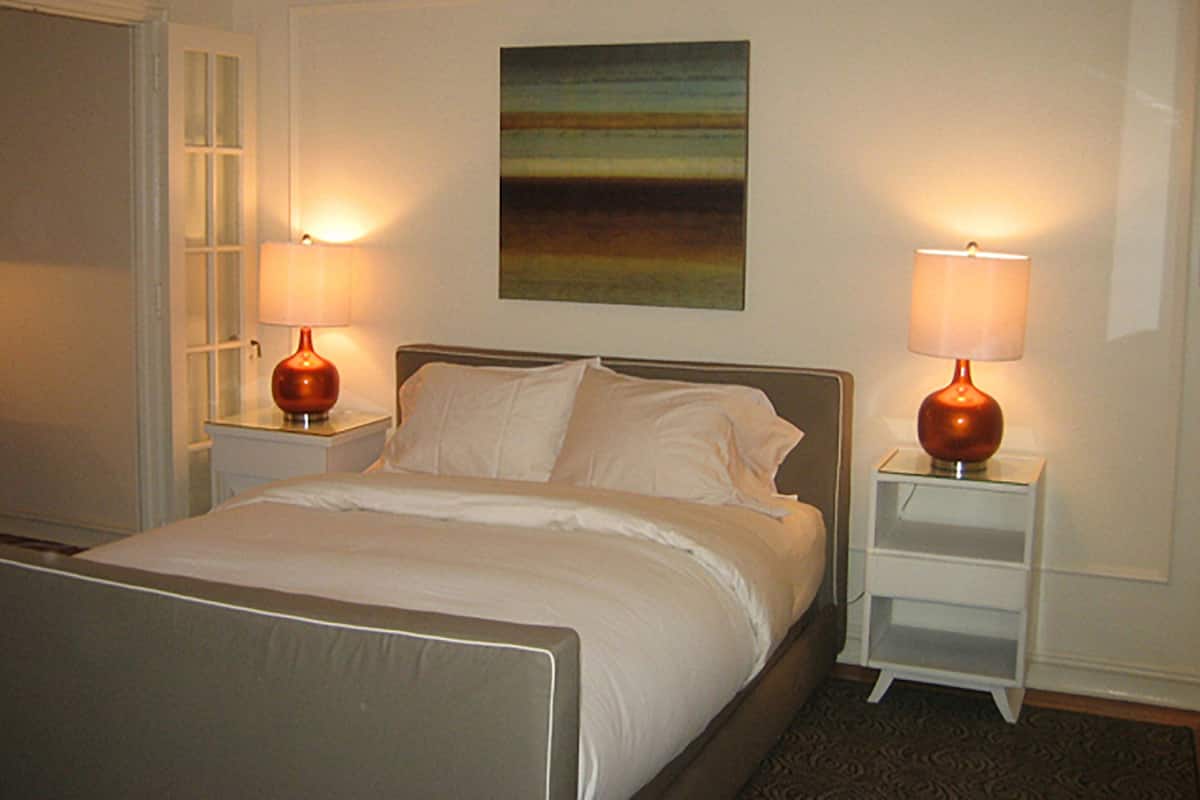 A neatly made bed with white linens, two bedside tables with lamps, a painting on the wall, and a chair in the corner.