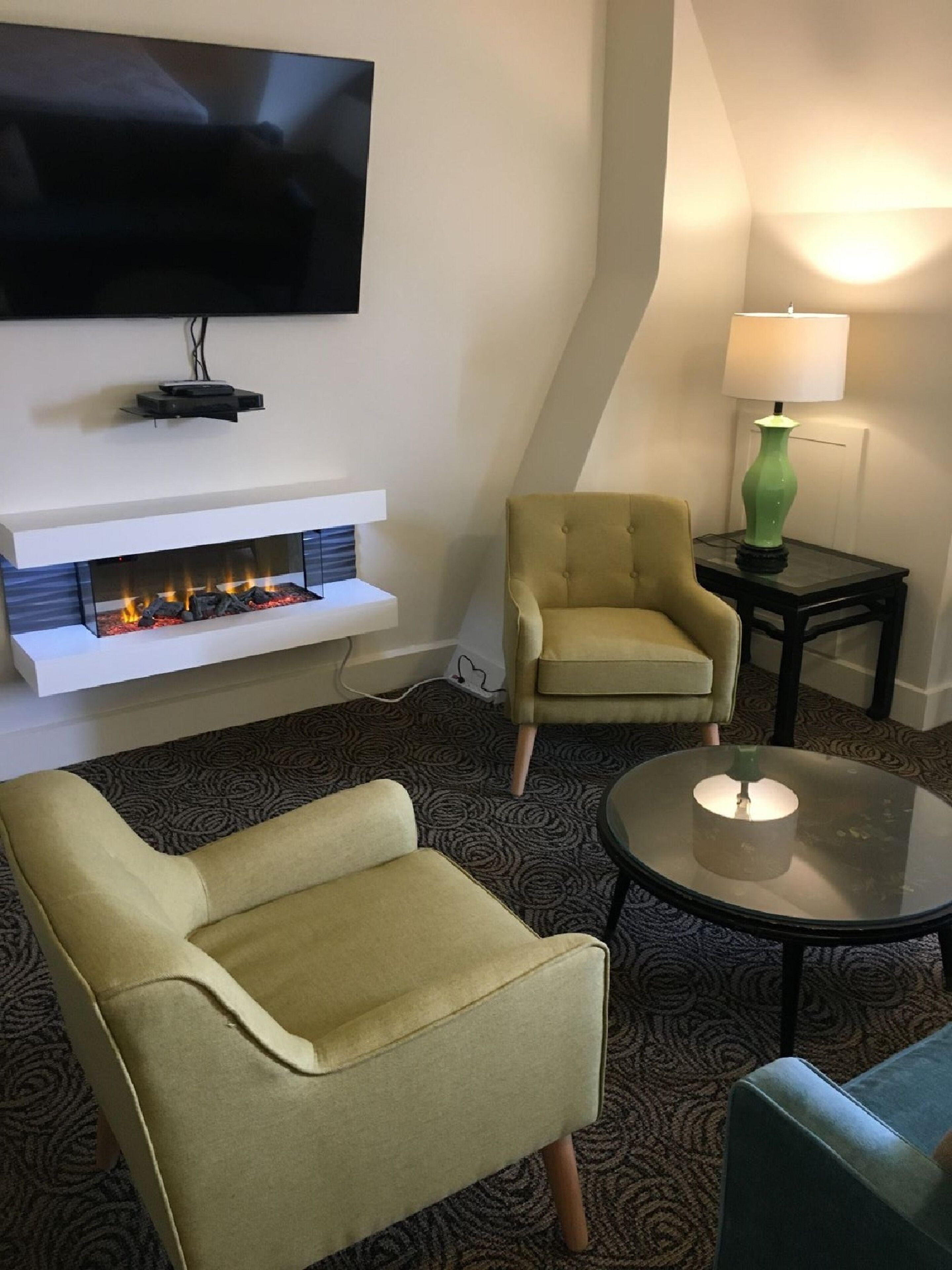 Ein Wohnzimmer mit flachbildfernseher an der Wand, einem Kamin mit modernem Design, einem grünen Sessel, einem runden Couchtisch mit Kerze und einer Lampe auf einem Beistelltisch.
