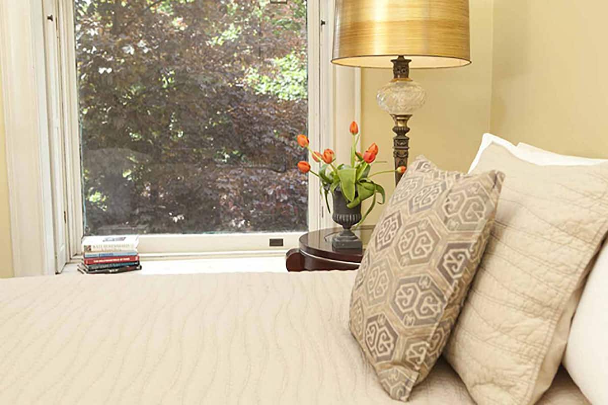 A neatly made bed with patterned pillows, a bedside lamp, a small table with a vase of flowers, and a window with a view of trees.