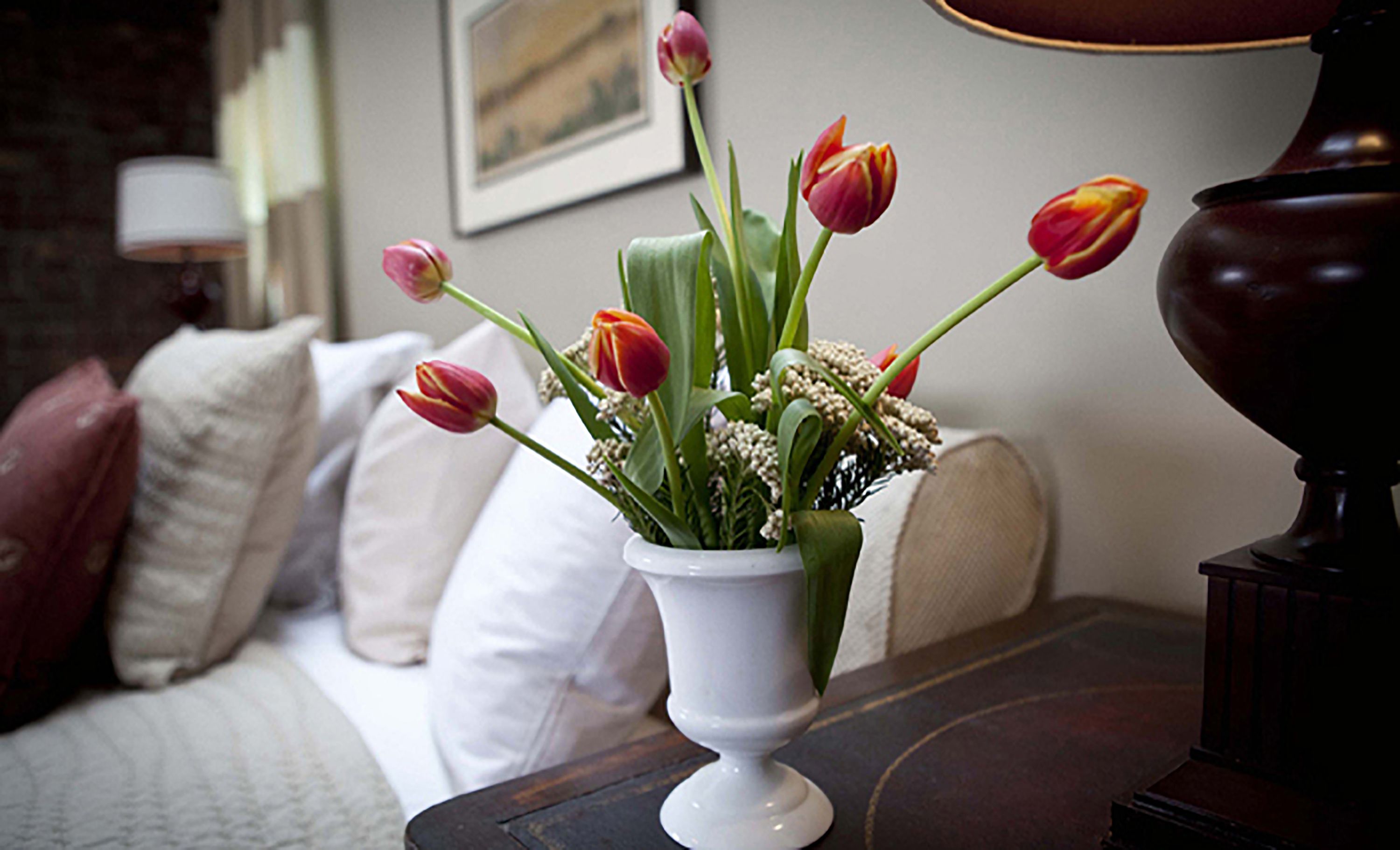 A white vase with tulips and baby's breath on a dark wooden table in a room with a white sofa and a lamp.