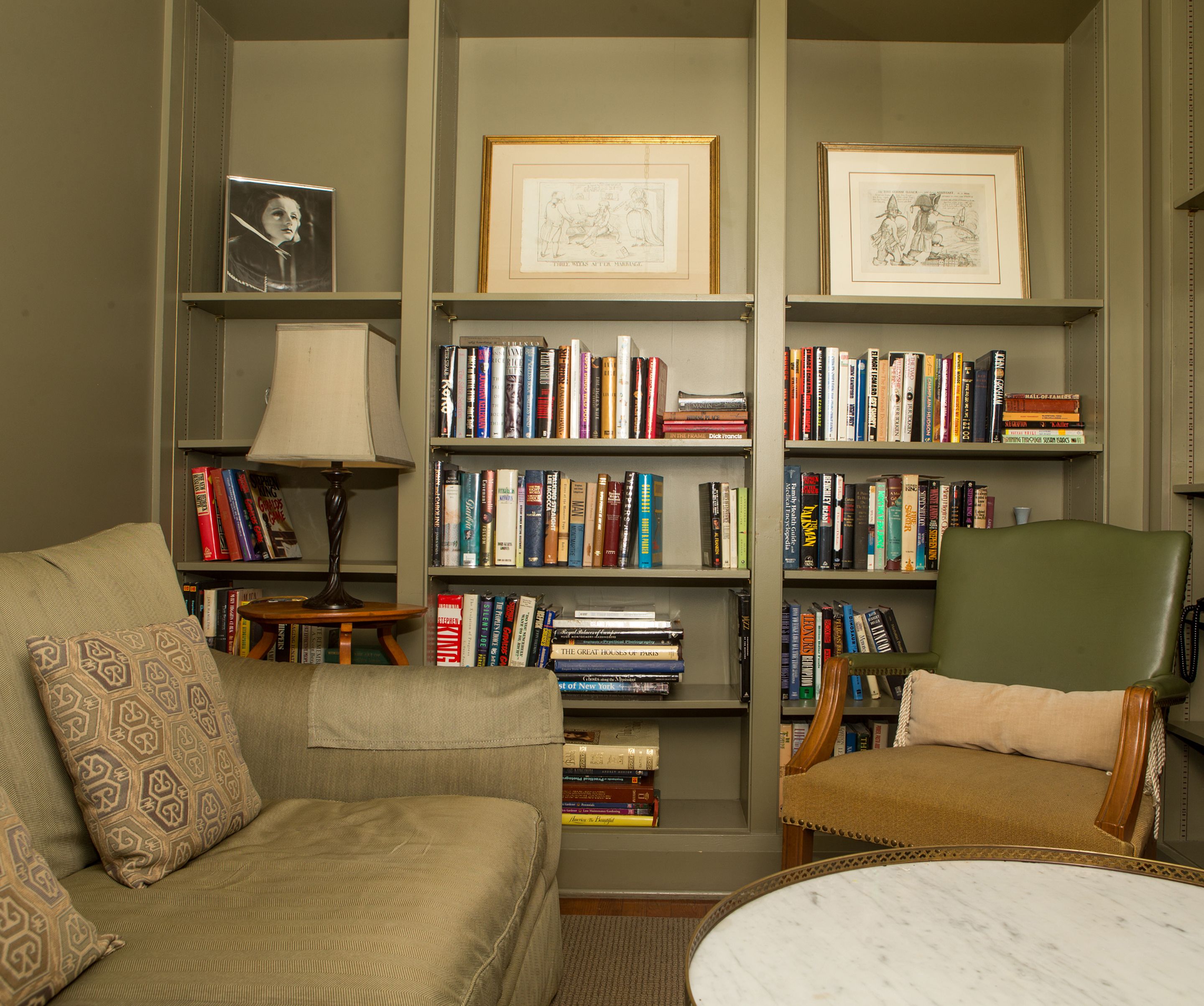 Un salon chaleureux avec un canapé, un fauteuil et une table basse. Des étagères remplies de livres et des tableaux encadrés ornent les murs.