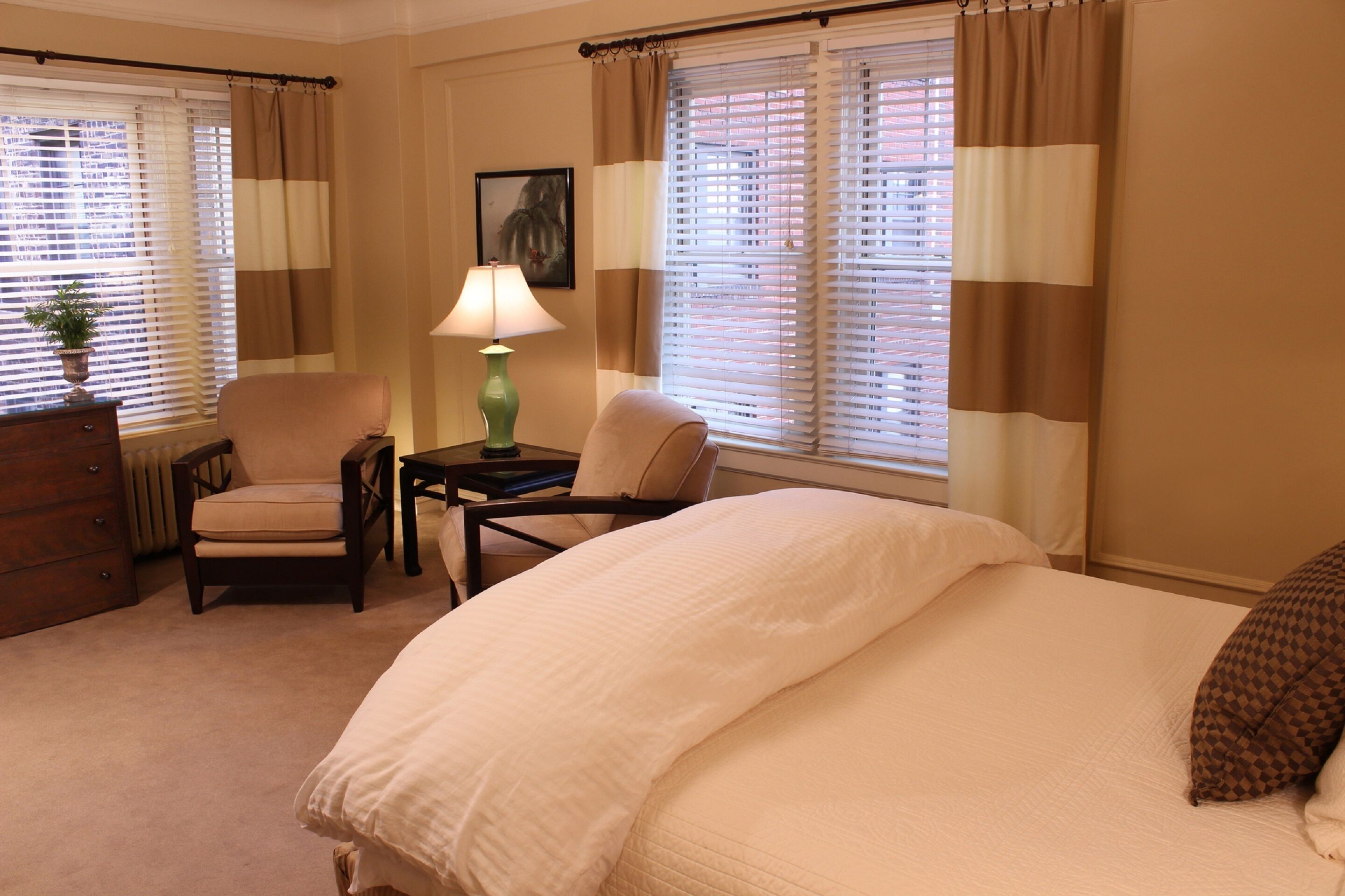 A hotel room with a large bed, two chairs, a dresser, a lamp, and windows with blinds.