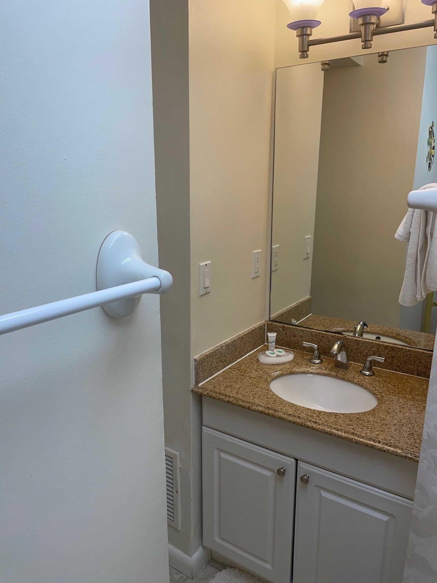 A bathroom with a white door, a towel rack, a sink with a granite countertop, a mirror, and a towel hanging on the wall.