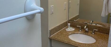 A bathroom with a white door, a towel rack, a sink with a granite countertop, a mirror, and a towel hanging on the wall.