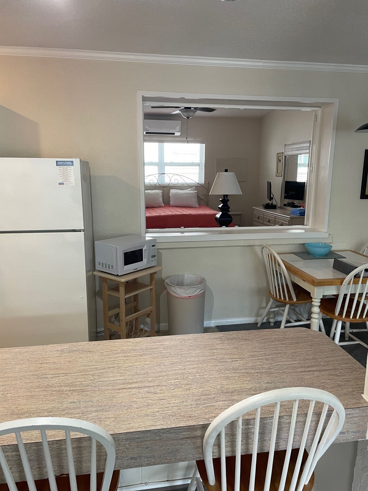 Une cuisine compacte équipée d’un réfrigérateur, d’un four à micro-ondes et d’une petite table à manger avec des chaises. Une fenêtre offre une vue sur un lit rouge et une lampe.